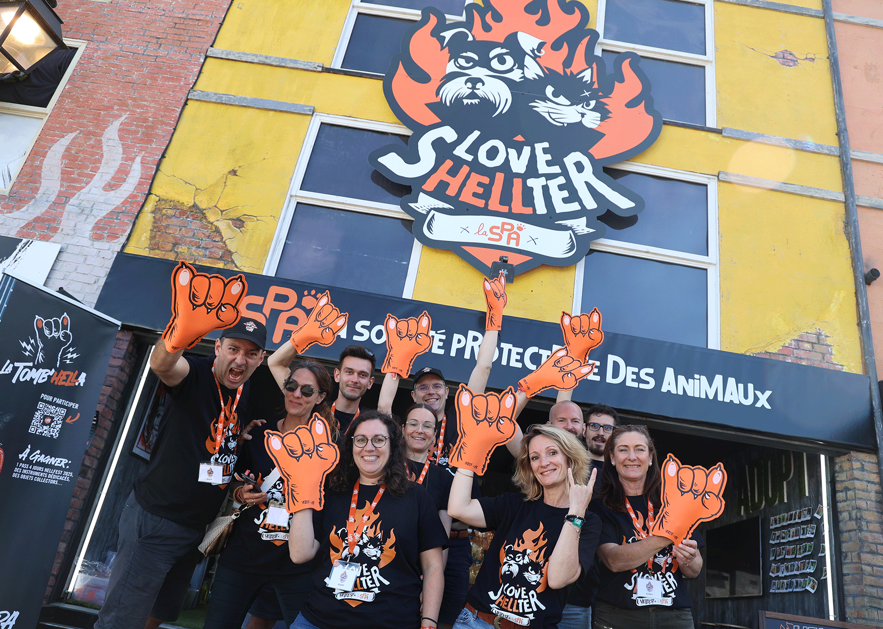 Les équipes de la SPA posant devant un stans du Hellfest 2024 avec une grande enseigne 'Love Shelter' et portant des gants en forme de patte de chat en mousse orange