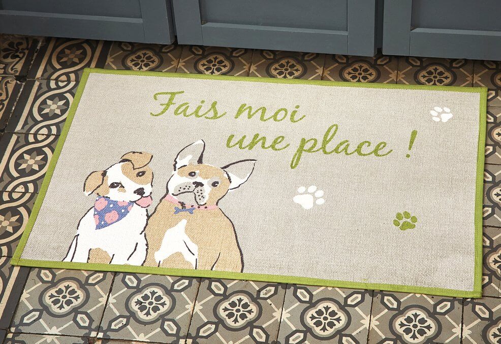 Un petit tapis de la marque Françoise Saget, avec un contour vert, on distingue 2 chiens et des empreintes de pas de chiens. C'est écrit : Fais moi une place !