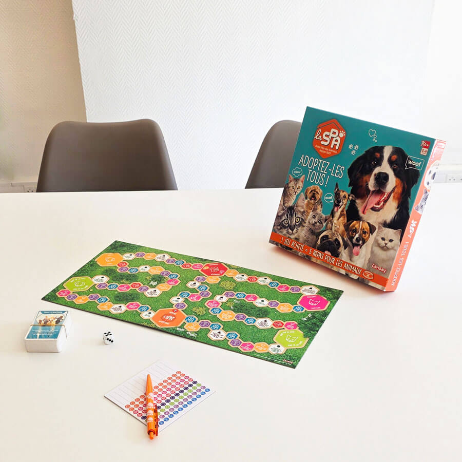 Un jeu de société sur le thème des animaux est disposé sur une grande table de réunion blanche. La boîte du jeu est à droite, montrant plusieurs chiens et chats de races diverses, avec le titre 'Adoptez-les Tous !' et le logo de la SPA. À gauche, le plateau de jeu déroulé représente un chemin en forme de spirale de cases hexagonales colorées sur un fond vert gazon. À côté du plateau, on trouve un dé, un petit bloc-notes blanc avec un stylo orange et des cartes à jouer. Deux chaises de bureau grises sont visibles à l'arrière-plan.
