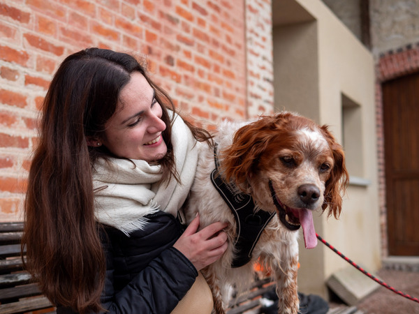 Une femme souriante caresse un chien roux et blanc tenu en laisse, devant un bâtiment en briques.
